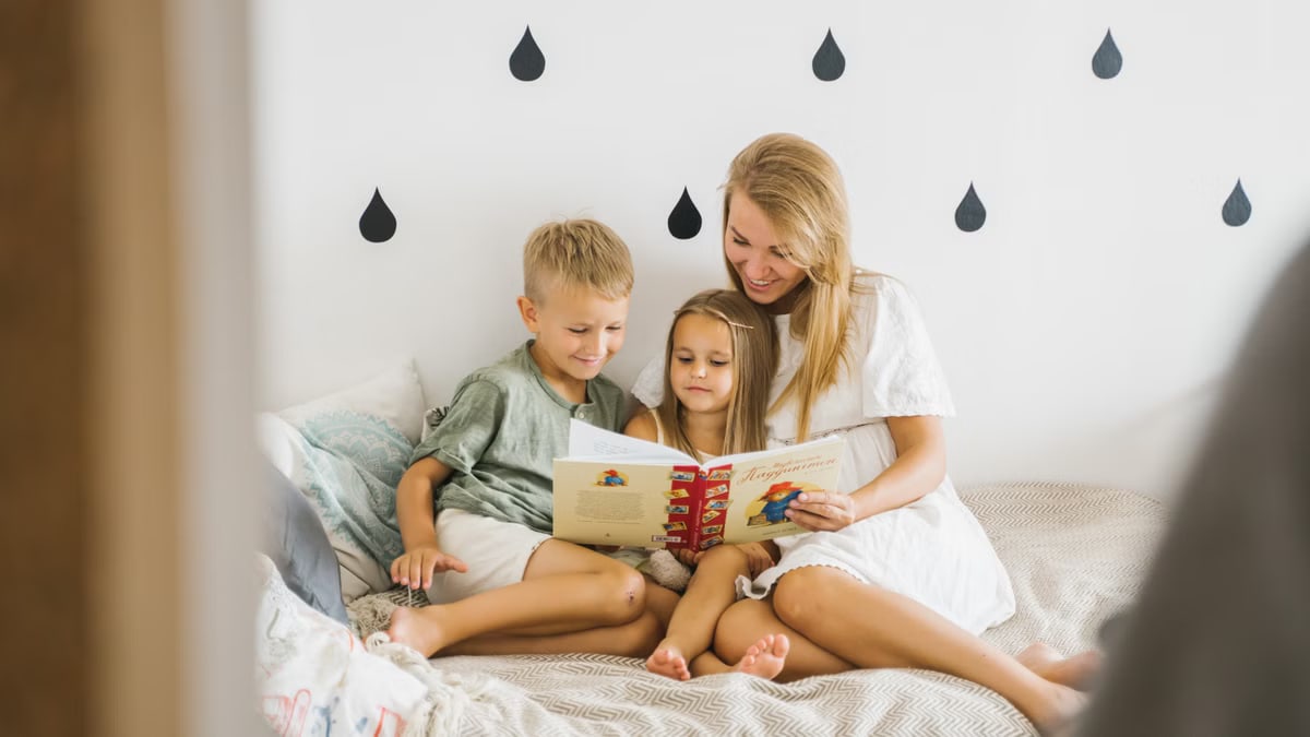 A joyful mother reads a storybook to her young children while sitting on a bed.